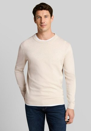 Man die een lichtbeige lange mouw trui draagt over een wit overhemd met donkerblauwe spijkerbroek, staand tegen een effen grijze achtergrond.
