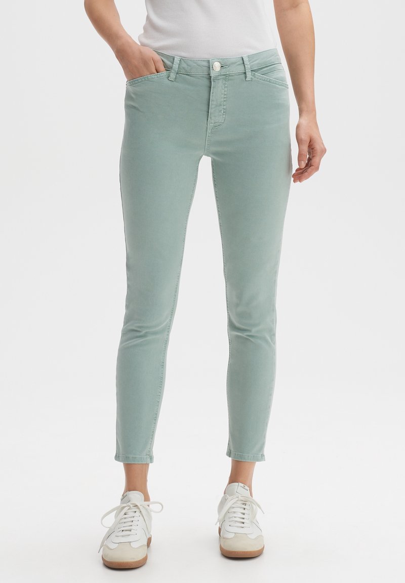 Personne portant un pantalon slim vert clair et des baskets blanches, debout avec une main dans la poche, sur un fond uni.