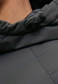 Chaqueta con capucha gris de un tejido texturizado, que presenta un sistema de ajuste en el cuello y un acabado liso y mate. Se aprecian detalles de costura.