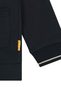 Felpa nera con una consistenza morbida, dotata di una tasca frontale, polsini a coste e un'etichetta gialla con il logo del marchio. Dettagli dorati sui polsini.