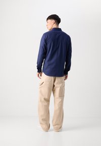 Cămașă bleu cu mânecă lungă și manșete cu nasturi, asortată cu pantaloni cargo bej largi, având buzunare laterale și o croială relaxată. Adidași albi vizibili.