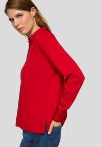 Roter, gestrickter Polo-Pullover mit langen Ärmeln, einem Stehkragen, seitlichen Schlitzen und einer lässigen Passform, kombiniert mit blauen Jeans.