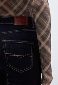 Vaqueros de mezclilla azul oscuro con costuras amarillas contrastantes, una etiqueta de cuero de la marca y una textura suave. Diseño visible del bolsillo trasero.