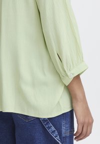 Blusa verde chiaro textured con maniche a tre quarti, caratterizzata da un piccolo bottone sul polsino e un orlo leggermente curvo.