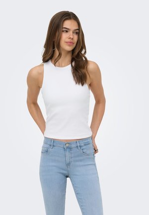 Jeune femme aux longs cheveux bruns portant un haut blanc sans manches et un jean bleu clair, debout les mains derrière le dos.