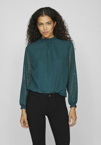 Blusa lunga teal con colletto arricciato, motivo a chevron testurizzato e polsini arricciati, abbinata a pantaloni neri aderenti.