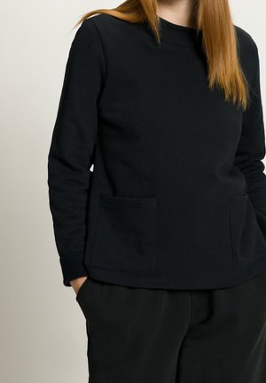 Femme portant un haut noir uni à manches longues avec deux poches à l'avant et un pantalon noir, debout avec une main dans la poche.