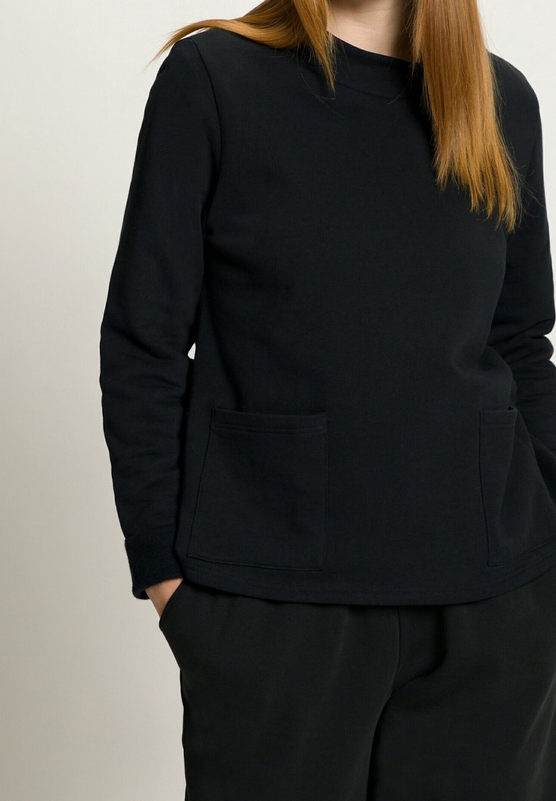 Femme portant un haut noir uni à manches longues avec deux poches à l'avant et un pantalon noir, debout avec une main dans la poche.