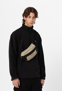 Borsa a vita beige e nera con una texture liscia, dotata di una cerniera superiore e di una tracolla indossata in diagonale. Il modello indossa un top in pile nero.