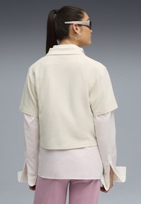 Kortärmad, ljusbeige texturerad topp med krage, lagd över en vit långärmad skjorta. Lavendelfärgade byxor syns nedanför.