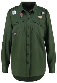 Camicia verde a maniche lunghe con due tasche sul petto, chiusura frontale con bottoni e vari toppe in tessuto tra cui un fenicottero e un cuore. Texture liscia.
