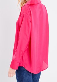 Blouse en satin rose vif avec col, manches longues et ourlet arrondi. Le tissu a une texture lisse et une finition brillante.