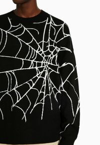 Svart tröja med ett vitt spindelvävs mönster, stickad textur, rund halsringning och kort skuren design med ribbade ärmslut och nederkant.