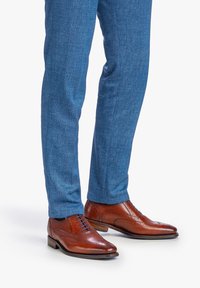 Scarpe brogue in pelle marrone con perforazioni intricate, design con lacci e punta arrotondata, abbinate a pantaloni blu texture.