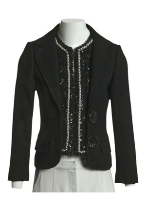 Blazer noir sur mesure avec deux gros boutons sur un top noir orné de sequins et de perles, associé à un pantalon blanc plissé sur un mannequin.