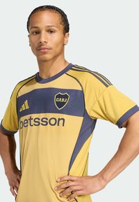 Gelbes und marineblaues Football-Trikot mit kurzen Ärmeln, ausgestattet mit dem "CABJ"-Logo, dem Text "betsson" und mesh-texturierten Akzenten auf der Brust.