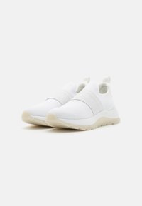 Chaussures blanches en maille à enfiler avec une surface texturée, dotées d'une large bande élastique au milieu du pied et d'une semelle de couleur claire.