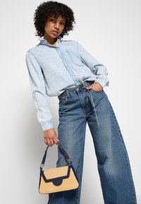 Camicia di jeans blu chiaro con bottoni e taschino; jeans blu a gamba larga; piccola borsa intrecciata gialla con dettagli blu marino e manico.