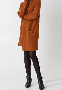 Robe-sweater orange brûlé avec manches longues, deux poches avant et coupe ample, associée à des bottines noires. Tissu doux et texturé.