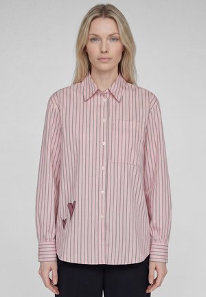 Blonde woman wearing a pink long-sleeve button-up shirt with thin black vertical stripes and two small heart designs near the waist.