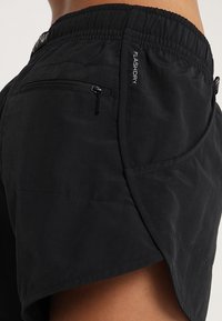 Närbild av svarta sportshorts med elastisk midja, bakficka med dragkedja, sidoficka och etikett med texten "FLASHDRY" på höger sida.