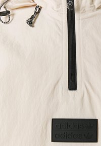 Ljus beige hooded jacka med halvdragkedja framtill, svart dragkedja, strukturerad tyg och en svart gummiyta med Adidas-logotyp.