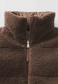 Braune Fleecejacke mit hohem Kragen, vollem Frontreißverschluss und einer strukturierten, plüschigen Oberfläche. Verfügt über einen markierten Reißverschlussgriff und ein Innenfutter.