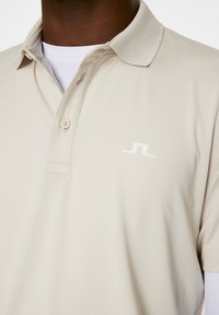 Beige pikétröja med kort ståkrage, med en enda knäppning och en vit logotyp på vänster bröst. Slät tygtextur.