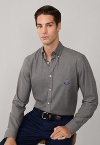 Hombre vistiendo una camisa gris de botones con logo, pantalones azul marino y cinturón trenzado, sentado contra un fondo gris claro liso.