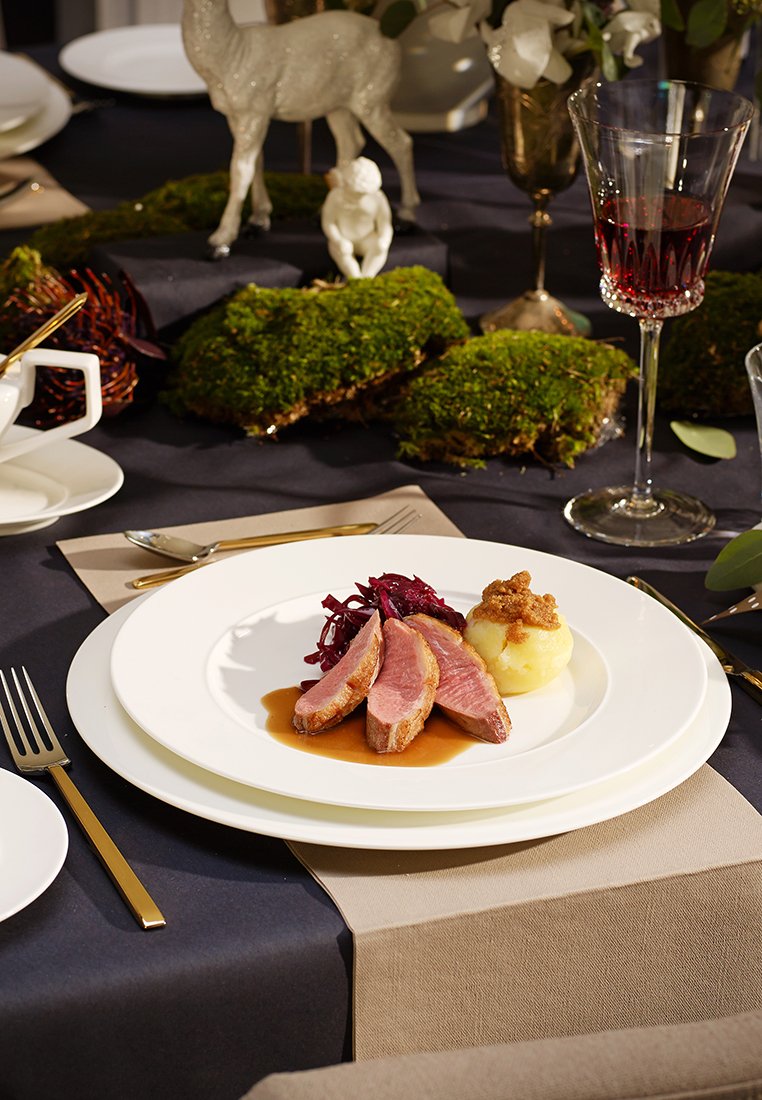 Ein Teller mit drei Scheiben rosa Entenbrust, Rotkohl und Kartoffelpüree mit brauner Soße. Weiße Keramik, goldverziertes Besteck, dunkle Tischdecke.