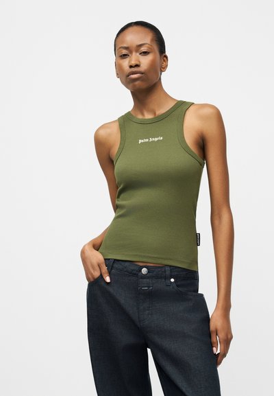 Slim woman wearing an olive green sleeveless top with "Palm Angels" text, paired with loose dark blue jeans, standing with one hand in pocket.
