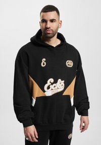 Hoodie noir avec cordon de serrage, présentant des panneaux diagonaux beiges et blancs, des accents de logo et une poche avant. Confectionné en tissu doux.