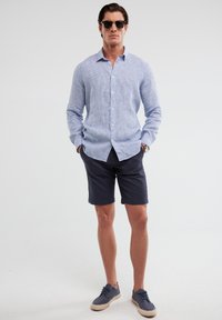 Camicia button-down a righe blu chiaro, pantaloni corti blu navy e sneakers grigie. La camicia ha un colletto classico e maniche lunghe.