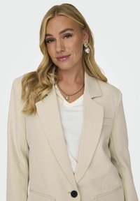 Blazer beige su misura con rever a punte, chiusura a un bottone e due tasche frontali, abbinato a una maglietta bianca a V.