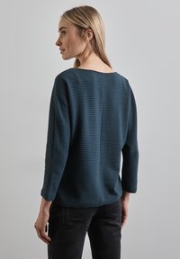 Tummansininen, kankainen neulepusero, jossa on leveä kaula-aukko ja kolmanneksen pituiset hihat. Ominaisuuksina suora leikkaus ja sivuhalkiot liikkuvuuden helpottamiseksi.