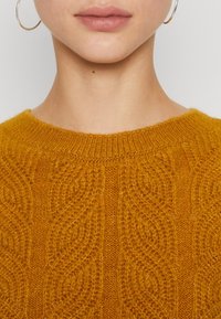 Nahaufnahme des unteren Gesichts einer Person, die goldene Creolen trägt und einen senfgelben, strukturierten Strickpullover mit einem runden Ausschnitt anhat.