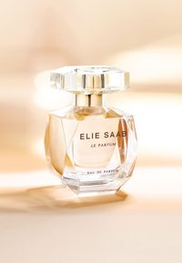 Klar glassparfymflaske med en diamantlignende form, en fasettert hette og gullkanter. Etiketten leser "ELIE SAAB LE PARFUM" med svart skrift.