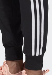 Svarta träningsbyxor med vit trestripesdesign på sidan. Muddarna är elastiska för en tät passform. Texturerad tyg, lämplig för aktivt bruk.
