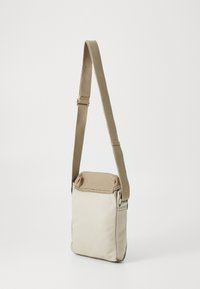 Bežová plátenná crossbody taška s hnedým vrchným klopovým vekom, nastaviteľným popruhom, obdlžníkovým tvarom a zipsovým uzáverom na boku.