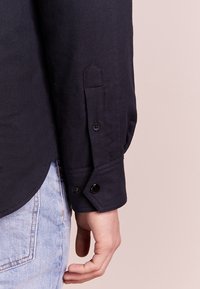 Camicia navy con collo strutturato; presenta polsini corti con bottoni neri e dettaglio a taglio triangolare. Tessuto in cotone testurizzato.