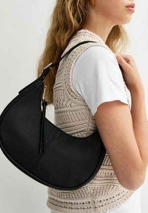 Mujer con cabello largo y rubio lleva camiseta blanca y chaleco de punto, cargando un bolso de hombro negro en forma de media luna con detalle de cremallera.