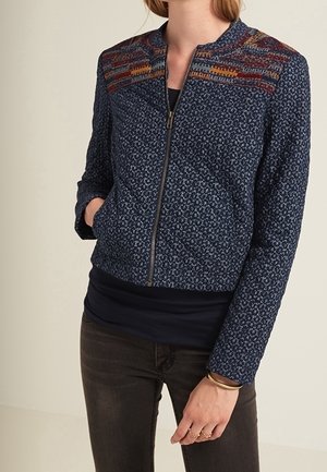 Kobieta w granatowej kurtce na suwak z wielokolorowym wzorem tkanym na ramionach, nosząca ciemne dżinsy oraz złotą biżuterię na nadgarstku i palcach.