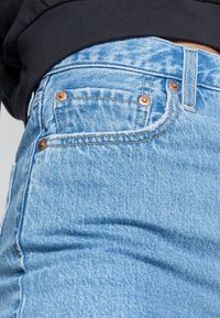 Los jeans de mezclilla azul claro presentan remaches de cobre, un bolsillo frontal y una silueta ajustada. La tela muestra un sutil patrón texturizado.