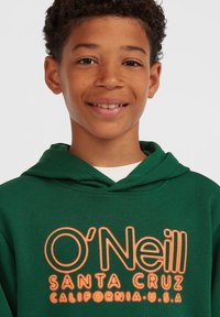 Smilende gutt iført en grønn hettegenser med "O'Neill Santa Cruz California U.S.A" trykket i oransje på forsiden, mot en ensfarget bakgrunn.