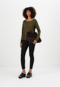 Top lungo a maniche lunghe verde oliva, pantaloni neri aderenti e sandali con platform neri. La modella tiene una borsa a tracolla morbida marrone scuro.
