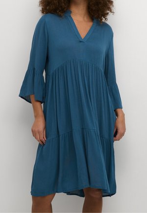 Nainen pukeutunut löysään, polvipituiseen siniseen mekkoon, jossa on V-pääntie ja kolmen neljänneksen hihat, seisoo yksiväristä taustaa vasten.