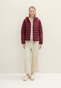 Chaqueta acolchada burdeos con capucha y cremallera frontal; presenta un diseño acolchado y puños elásticos. La modelo lleva pantalones anchos de pierna en beige claro y zuecos.