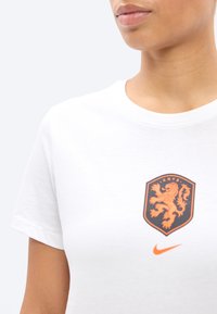 Personne portant une chemise blanche avec un emblème de lion orange et le texte "KNVB" au-dessus du logo Nike swoosh sur la poitrine.