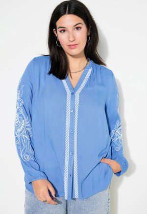 Blusa azul clara con mangas largas, que presenta bordados blancos en las mangas y un cierre de botón en la parte delantera. Tejido texturizado.