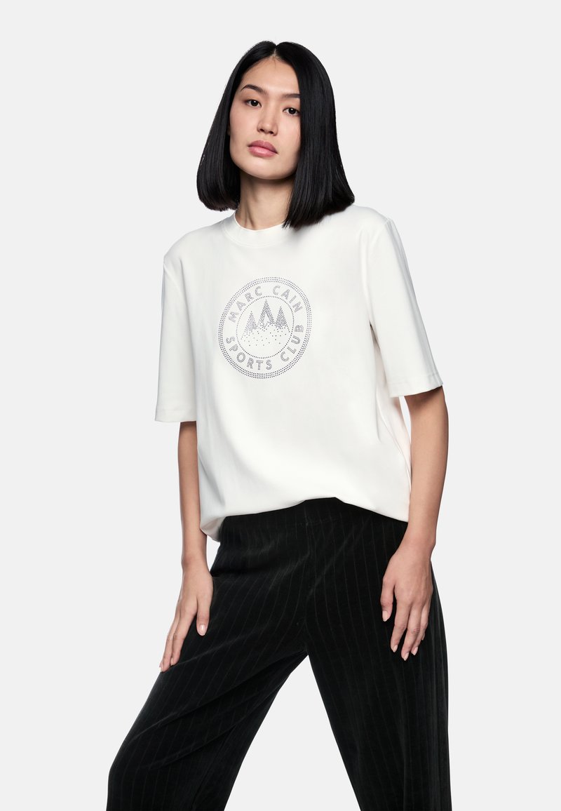 White short-sleeve t-shirt with a circular graphic in gray featuring mountain and text detail. Smooth fabric, relaxed fit. Black striped pants.